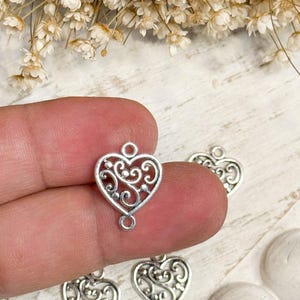 May include: A silver-toned metal heart charm featuring an intricate filigree scrollwork pattern. The heart-shaped charm includes a small circular loop at both the top and the bottom, designed for connecting to other jewelry components or chains.