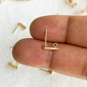 May include: Gold earring findings with a small loop and a straight post. The findings are shown on a finger.