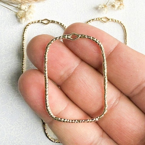 May include: Close-up of several gold-colored rectangular hoop earring components. The hoops have a textured surface and a small, diamond-shaped connector at the top. The background includes dried flowers and twine.