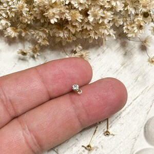May include: A small, gold-colored stud earring with a clear, round gemstone. The earring is held between fingers, with a blurred background of dried flowers and a light-colored surface.