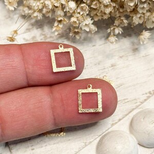 May include: Two small, gold-colored square charms with a textured, matte finish. Each square features a hollow center and a small circular loop at the top for attachment. The charms are held in place, showcasing their geometric shape and metallic surface.