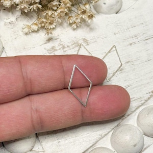 May include: A pair of silver-colored diamond-shaped earrings. One earring is held between fingers, while another rests nearby. The earrings are simple geometric shapes, suitable for everyday wear. The background includes dried flowers and decorative spheres.