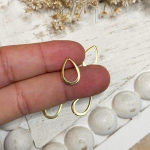 May include: A small, gold-colored teardrop-shaped earring held between fingers, with a larger matching earring in the background. The earrings are simple and elegant, with a minimalist design. The background includes dried flowers and a textured surface.