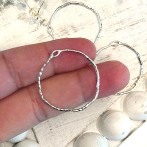 May include: Silver-toned, hammered metal hoop earrings with an irregular, textured edge. The earrings are circular with a small loop at the top for attachment. The image shows several earrings, with one held in a hand.