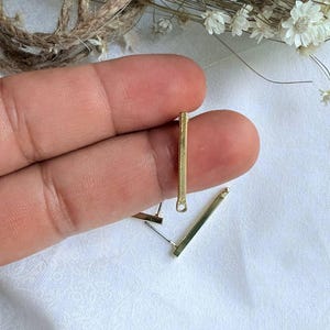 May include: Two gold bar earrings with a loop at the top. The earrings are shown on a finger.