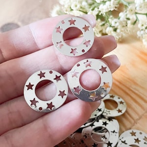 May include: Silver-colored metal circle charms with star cutouts. The charms have a circular hole in the center and are arranged in a hand. The background includes white flowers.