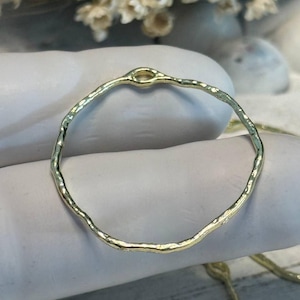 May include: A gold-colored, hammered metal ring with an irregular, organic shape. The ring has a small loop at the top and a textured surface. The ring is likely a jewelry component or a decorative element.