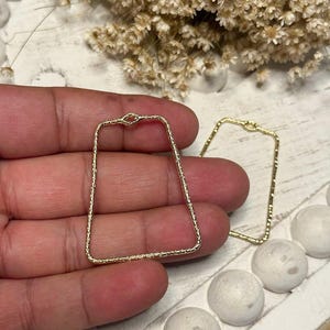 May include: Two gold-colored, rectangular-shaped earring components. The earrings have a textured, beaded appearance. The top of the earrings have a small, decorative loop. The earrings are displayed on a white surface with decorative elements.
