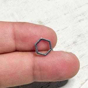 May include: A small, silver-colored, open-frame hexagon shape. The geometric design is held between two fingers against a white and light wood grain background. The hexagon is a simple, modern design.