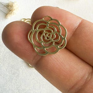 May include: Gold metal rose charm with a delicate, intricate design. The charm is a cutout shape, with a circular outline and a detailed rose pattern.