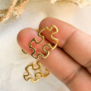 May include: Two gold-colored puzzle piece charms. The charms are made of metal and have a cutout design.