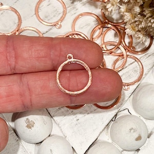 May include: A close-up of a rose gold metal pendant with a circular frame. The pendant is being held by a finger. There are several other similar pendants in the background.