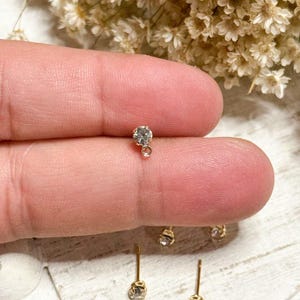May include: Close-up of gold-colored stud earrings with a small, clear gemstone. The earrings have a simple design with a single, round stone. Several pairs of earrings are visible, along with a finger holding one earring. The background features dried flowers.