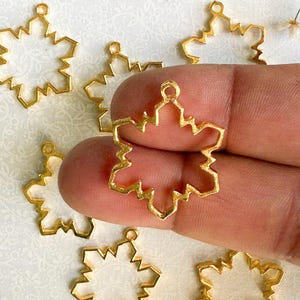 May include: Six gold-tone metal snowflake-shaped pendant blanks. The blanks are open and have a smooth, shiny finish.