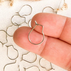 May include: A close-up of a silver earring finding, a type of jewelry component used to attach earrings to a hook or post. The finding is shaped like a small hoop with a slight bend at the top. There are multiple findings scattered on a white surface.