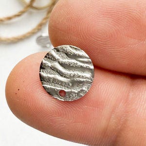 May include: A silver round charm with a textured surface and a small hole in the center.