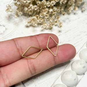 May include: Two gold-colored, open-frame diamond-shaped earrings. The earrings are displayed against a background of dried flowers and white decorative spheres. The earrings are small and delicate, suitable for everyday wear.