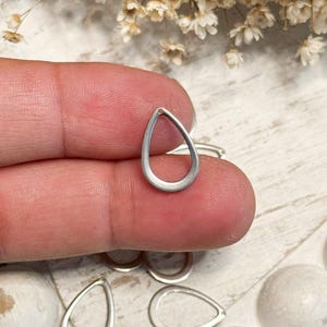 May include: A small, silver-toned metal charm in the shape of a teardrop or open pear outline. The charm features a smooth, matte finish and a small hole at the top point for attachment. Several identical teardrop-shaped metal charms are visible.