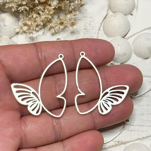 May include: Two silver-colored butterfly wing-shaped earring charms. Each charm features a delicate openwork design with a teardrop shape and detailed wing patterns. The charms are shown against a soft, neutral background.
