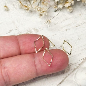 May include: Close-up of small, gold-colored diamond-shaped charms. The charms have a textured surface and are displayed on a person's finger. White flowers and decorative stones are in the background.