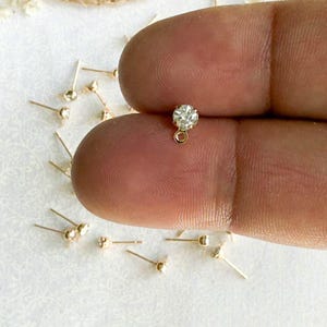 May include: Close-up of a small, gold-colored earring with a clear, round gemstone. The earring is held between fingers, with several other similar earrings scattered on a white surface. The earrings have a post and a small loop.