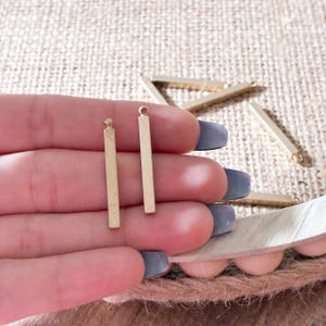 May include: Close-up of gold-colored rectangular bar charms. Two charms are held in a hand, with several more scattered in the background. The charms have a small loop at the top for attaching to jewelry. The background is a neutral tan color.