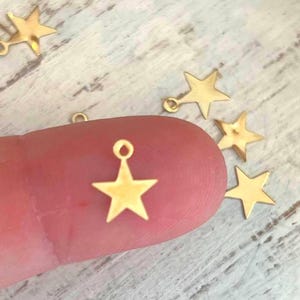 May include: Gold star charm with a loop at the top. The star is a simple design with five points. The charm is on a finger.