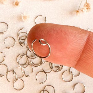 May include: A close-up of a silver nose ring on a finger. The ring is a simple hoop with a small bead on the top. There are many other nose rings scattered around the ring on the finger.