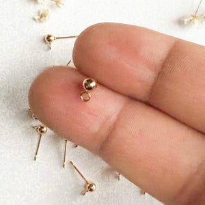 May include: Gold-toned earring findings with a small ball at the end of each post. The findings are shown on a finger.