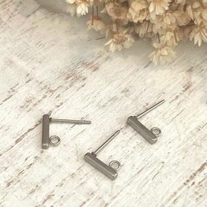 May include: Three silver-toned earring posts with a rectangular bar and a loop for attaching charms. The posts are on a white, distressed wooden surface, with dried flowers in the background. The earring findings are for jewelry making.