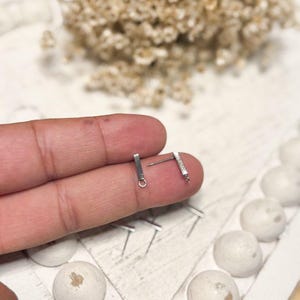 May include: Two silver bar earrings with a small loop and post. The earrings are held by a person's finger. The background includes small white spheres and dried flowers on a white surface.