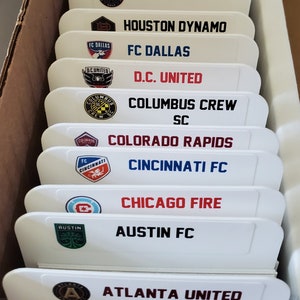 May include: A white shelf with labels for different Major League Soccer teams. The labels feature the team's logo and name. The teams include LA Galaxy, Inter Miami CF, Houston Dynamo, FC Dallas, D.C. United, Columbus Crew SC, Colorado Rapids, Cincinnati FC, Chicago Fire, Austin FC, and Atlanta United.