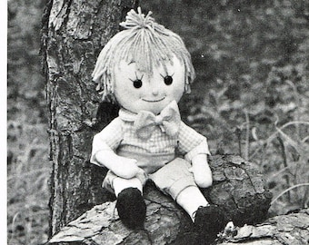 May include: A handmade rag doll with brown hair, wearing a plaid shirt and a bow tie, sitting on a log.