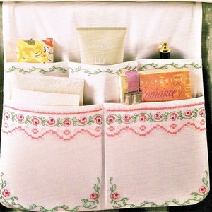 May include: White fabric organizer with six pockets, embroidered with pink flowers and green vines. The organizer is filled with various items, including a book titled "Arlequin Romance", a tube of hand and body lotion, and a small white towel.