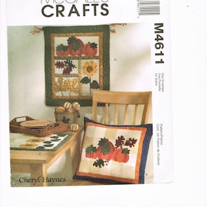 Op de afbeelding: Een McCall's Crafts quiltpatroon voor een herfst-thema kussen en wandhanger. Het kussen heeft een ontwerp van pompoenen en bladeren, terwijl de wandhanger een vergelijkbaar ontwerp heeft met de toevoeging van een zonnebloem. Het patroon is getiteld "M4611" en is van Cheryl Haynes.