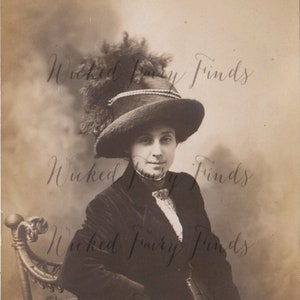 May include: A black and white vintage photograph of a woman wearing a dark hat with a feather and a dark coat. She is sitting in a chair with a decorative back.