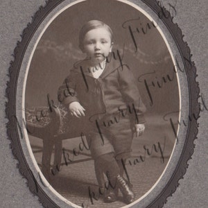 May include: Sepia-toned portrait of a young child in a dark suit and shorts, seated on a small chair. The child wears dark boots and a sailor-style jacket. The image is framed within an oval border.