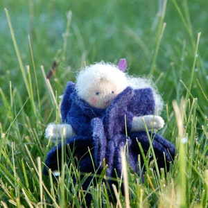 May include: A handmade doll with a knitted face, white hair, and a blue felt outfit. The doll is sitting in a patch of green grass, with a purple accent on its head. The doll's arms are made of a light beige material.