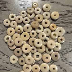 May include: A pile of off-white donut-shaped beads with a hole in the center. The beads are made of bone or wood and have a natural, rustic look.
