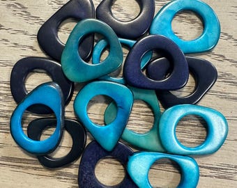 May include: A collection of teardrop-shaped, open-center beads in shades of blue and teal. The beads vary in color from deep navy to bright turquoise, with some showing a marbled effect. The collection is arranged on a light-colored, wood-grain surface.