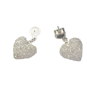 Puede incluir: Un par de pendientes de plata en forma de corazón con un diseño delicado e intrincado. Los pendientes son de estilo colgante y tienen un cierre de poste.