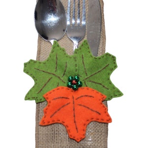 May include: A burlap napkin holder with a green and orange felt leaf design. The holder has a spoon, fork, and knife inside.