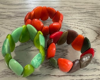 Pode incluir: Três pulseiras elásticas compostas por contas lisas, arredondadas e em forma de seixo. Uma pulseira é laranja sólida, uma tem vários tons de verde e a terceira apresenta uma mistura de contas laranja, castanhas, verde-escuras e cinzentas.