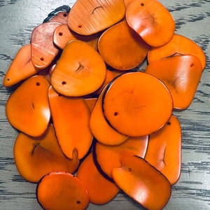 May include: A collection of orange, irregularly shaped, polished, natural shell beads with holes drilled through them. The beads are arranged in a pile on a gray surface.