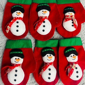 May include: Six red felt mittens with green cuffs, each featuring a white snowman with black eyes, a black hat, and a red scarf with white polka dots.