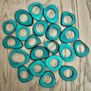 May include: A collection of teal-colored, irregularly shaped pendants with a hole in the center. The pendants are arranged on a light-colored wooden surface. Each pendant has a small hole at the top, suggesting they are designed for jewelry making or crafts.