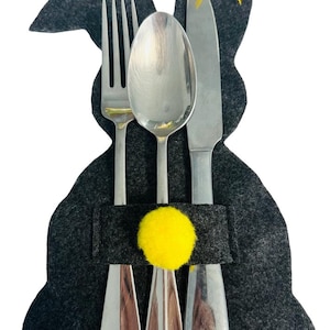 May include: A gray felt bunny-shaped cutlery holder with a yellow pom-pom and a yellow ribbon bow. A fork, spoon, and knife are placed inside the holder.