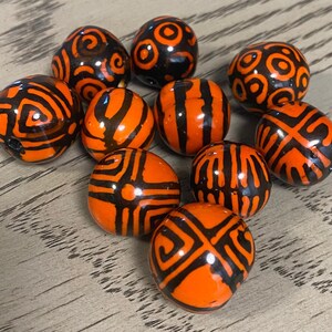 May include: A collection of orange and black patterned beads. The beads have various designs, including swirls, stripes, and geometric shapes. The beads are round and appear to be made of a glossy material.