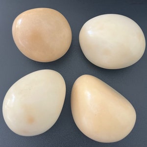 Può includere: Quattro pietre lisce di forma ovale in tonalità crema e marrone chiaro. Le pietre sono disposte a forma di quadrato su una superficie grigio scuro.