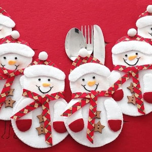 May include: Set of six white felt snowman napkin rings with red and gold trim. Each snowman has a red hat, a red scarf, and a red button nose. The snowmen are arranged around a silver fork and spoon.